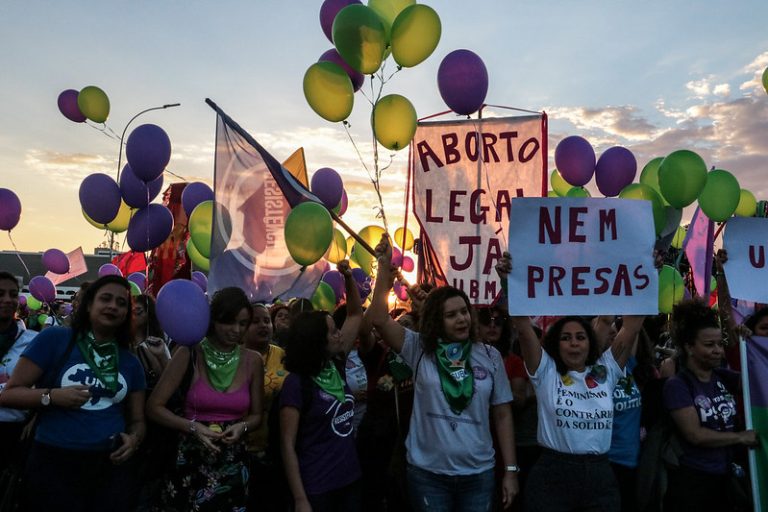 Mulheres em manifestação com balões verdes e roxos, seguram cartazes onde se lê "aborto legal já".