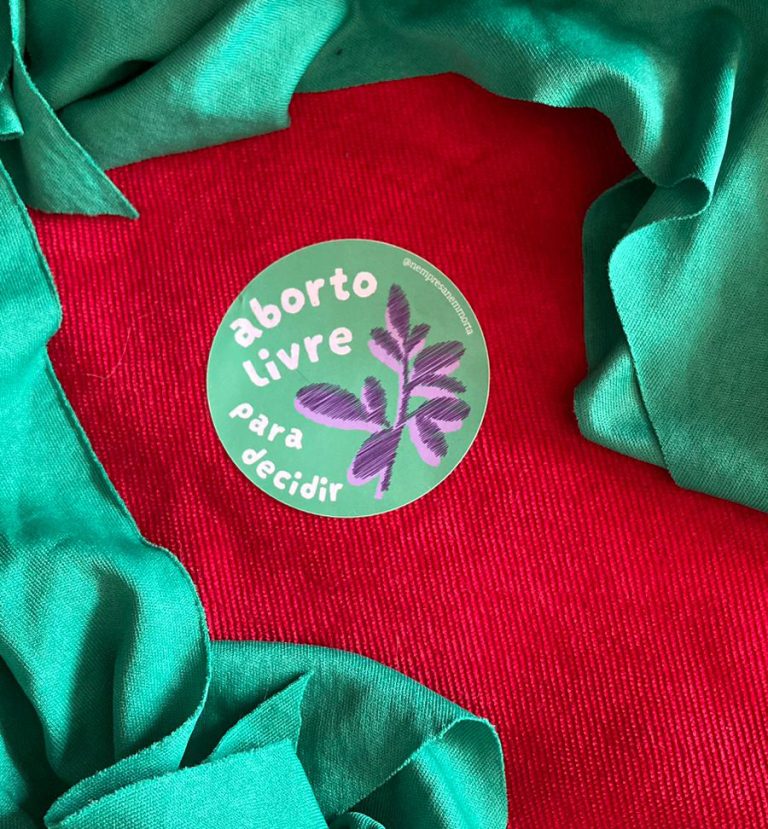 Fundo vermelho e verde. Adesivo no centro. O adesivo é redondo, com fundo verde. Em branco lê-se aborto livre para decidir. ao lado há uma ilustração da folha de arruda em roxo e verde.