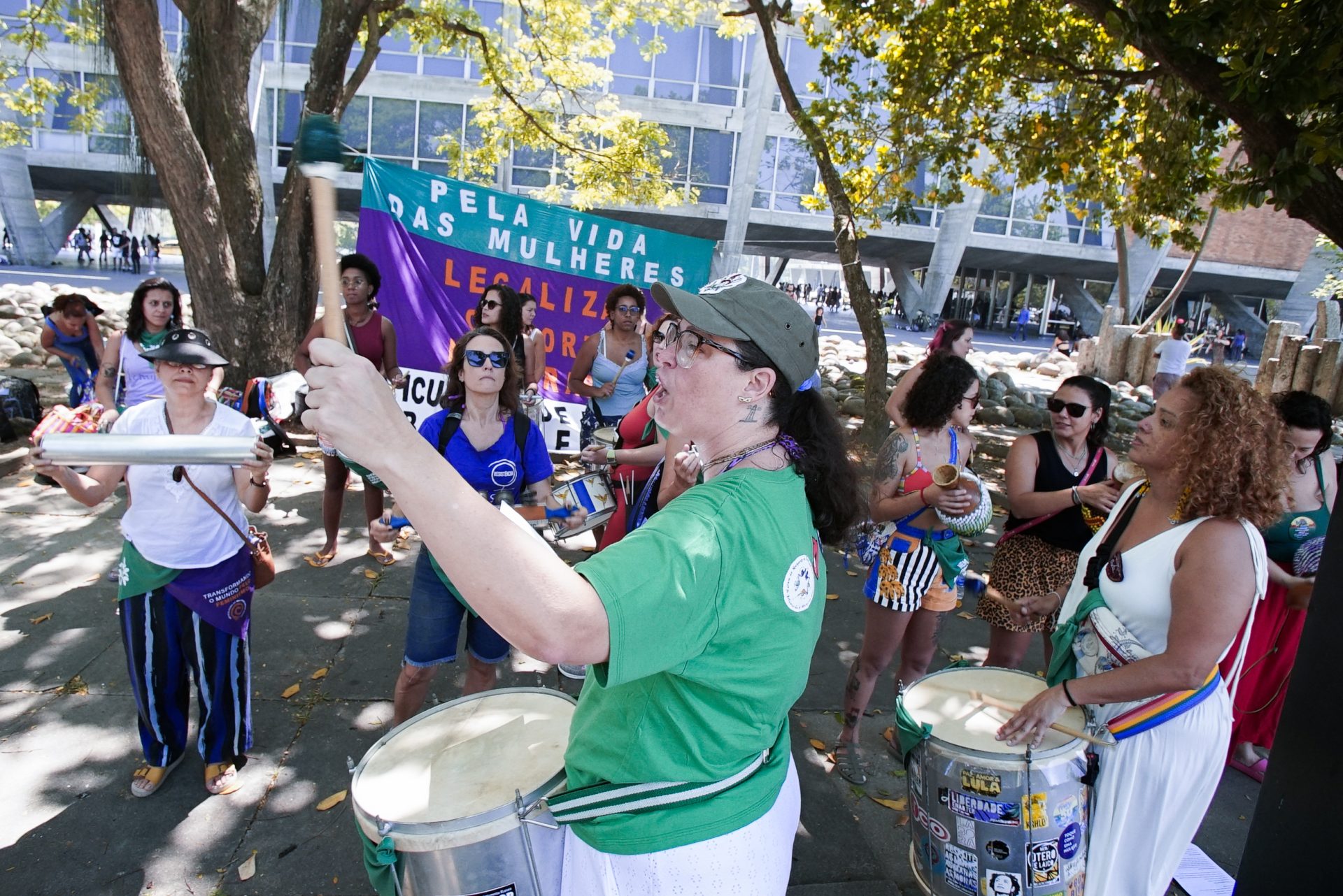 Diversas mulheres tocam instrumentos e batucada. Atrás há uma bandeira em roxo e verde.