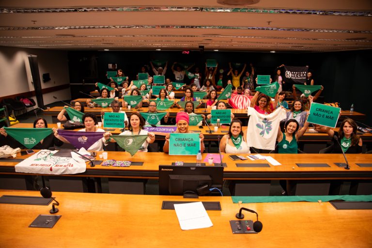 Diversas mulheres estão sentadas no plenário da Câmara Federal segurando lenços e cartazes pela descriminalização do aborto na América Latina e Caribe.