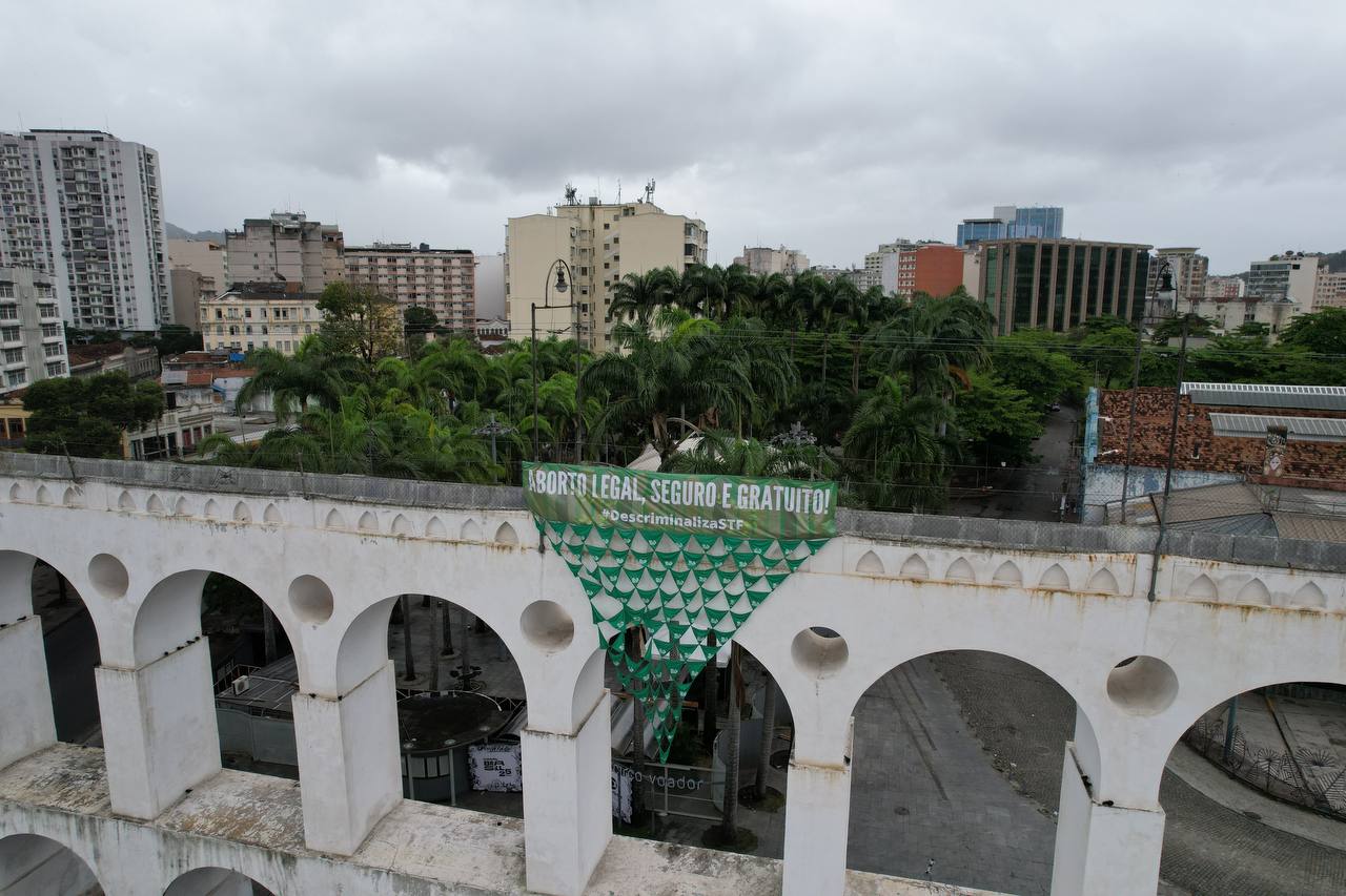 Vista aérea dos arcos da lapa, no Rio de janeiro. Há uma faixa verde que diz "Aborto legal, seguro e gratuito. Descriminaliza STF". Abaixo há uma bandeira formada por diversos lenços unidos pelas pontas, que formam um triângulo.
