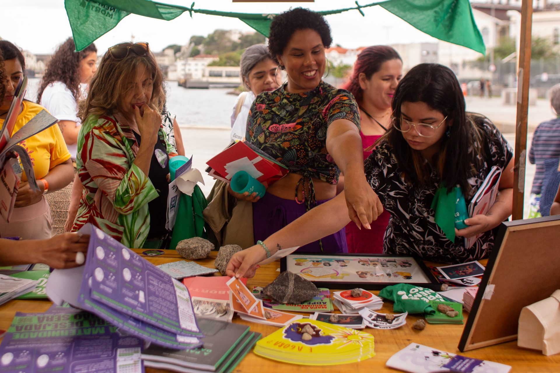 Foto de uma mesa, que fazia parte da barraca de Justiça Reprodutiva, onde á materiais diversos dispostos. Algumas mulheres observam e pegam os materiais que estão sobre a mesa.