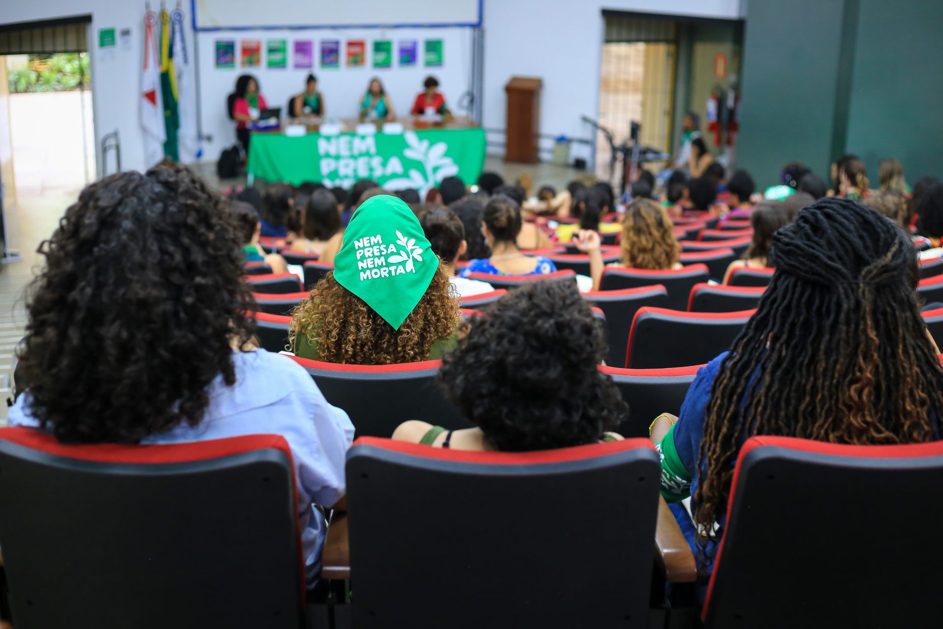 Imagem de um auditório. Vemos as costas de mulheres sentadas e, ao fundo, uma mesa coberta com um lenço verde da Campanha Nem Presa Nem Morta onde estão sentadas quatro mulheres palestrantes.