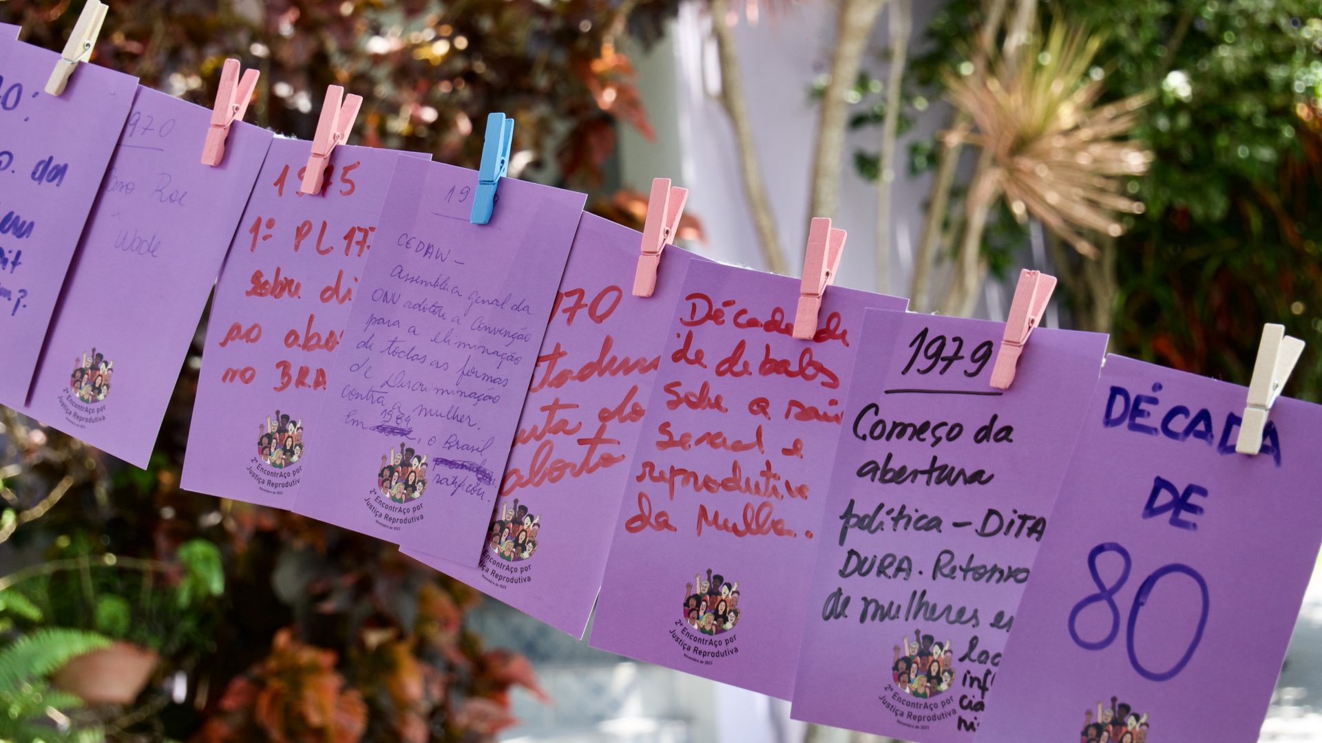 Varal com papéis lilás e prendedores rosas, amarelos e azuis. os papéis têm mensagens que resgatam a história da luta pelo direito ao aborto.