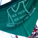 Ienço verde pendurado, com ilustração de uma mulher e dizendo: "nem presa, nem morta, aborto livre para decidir".