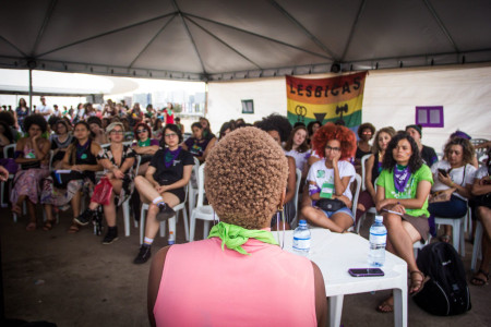 Foto: Mídia Ninja. Plateia ouve uma mulher negra de cabelos curtos, que aparece em primeiro plano, de costas na foto.