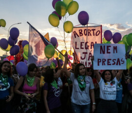 Foto: Chama. Mulheres em manifestação com balões verdes e roxos, seguram cartazes onde se lê "aborto legal já".