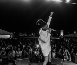 Foto: Mídia Ninja. Foto de cima de um palco. Mulher negra com o braço erguido canta para uma planteia. Foto em preto e branco.