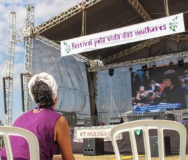 Foto: Chama. Em primeiro plano, uma mulher sentada, com camiseta roxa, observa um palco com uma faixa onde se lê "Festival pela vida das mulheres".