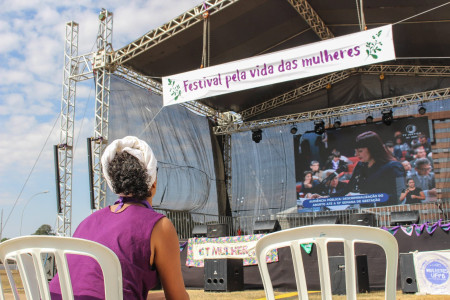 Foto: Chama. Em primeiro plano, uma mulher sentada, com camiseta roxa, observa um palco com uma faixa onde se lê "Festival pela vida das mulheres".