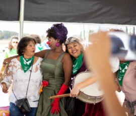 Foto: Chama. Três mulheres se abraçam, a do meio está caracterizada como palhaça.