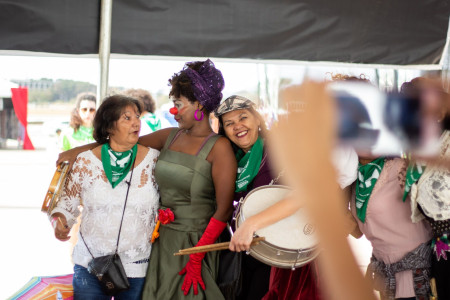 Foto: Chama. Três mulheres se abraçam, a do meio está caracterizada como palhaça.