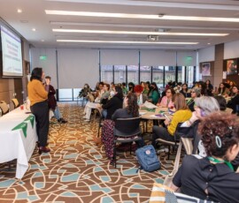 Sala de reuniões. Há uma mulher de amarelo e preto na frente, as demais estão em mesas circulares, divididas em grupo.