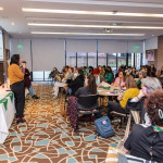 Foto de Monica Cotes - Trineo Comunicaciones. Sala de reuniões. Há uma mulher de amarelo e preto na frente, as demais estão em mesas circulares, divididas em grupo.