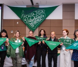 Sete pessoas, sendo 6 mulheres e um homem, seguram lenços verdes e posam em frente a um grande lenço onde se lê "De Causa Justa la Libertad es imparable", em espanhol.