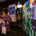 Foto de Monica Cotes - Trineo Comunicaciones. Mulher de casaco vermelho quadriculado fotografa painel que expõe lenços verdes e cartazes da campanha Causa Justa.