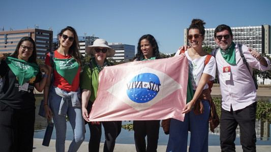 Duas mulheres negras, três mulheres brancas e um homem branco seguram uma bandeira "Viva o SUS", que imita a bandeira do Brasil, mas nas cores rosa, azul e branco. Algumas pessoas vestem lenços verdes e óculos escuros.