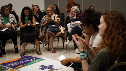 Mulheres sentadas em roda, sorrindo e batendo palmas. Ao centro vê-se bandeiras de movimentos sociais e políticos.