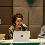 Foto de Diana Ramirez. Mulher branca sentada atrás de um computador falando ao microfone.