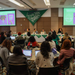 Foto de Diana Ramirez. Sala de reunião. Visão de uma mesa de debatedoras ao fundo e participantes em mesas circulares em frente.
