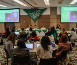 Sala de reunião. Visão de uma mesa de debatedoras ao fundo e participantes em mesas circulares em frente.