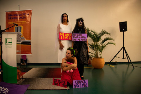 Três mulheres posam com cartazes e roupas de performance.