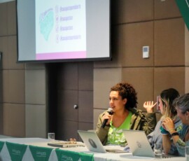 Mesa de debatedoras, onde vê-se três mulheres apresentando slides ao fundo.