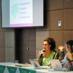 Foto de Diana Ramirez. Mesa de debatedoras, onde vê-se três mulheres apresentando slides ao fundo.