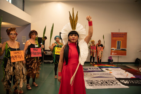 no centro há uma mulher indígena vestida de vermelho e mulheres com roupas de performance a rodeiam.