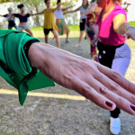 Mão com lenço verde amarrado no pulso. Em segundo plano, ao fundo mulheres em pose de yoga.