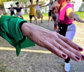 Mão com lenço verde amarrado no pulso. Em segundo plano, ao fundo mulheres em pose de yoga.