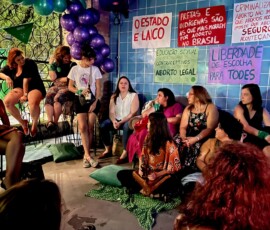 Mulheres em roda conversam. Atrás uma parece azul com diversos cartazes.