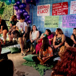 Mulheres em roda conversam. Atrás uma parece azul com diversos cartazes.