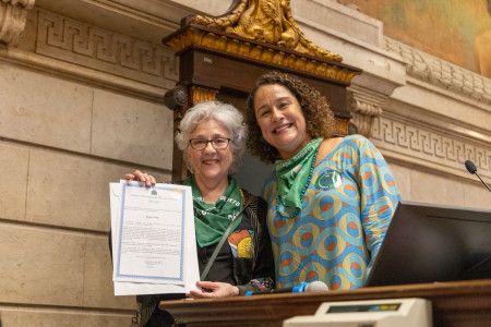 Angela Freitas recebeu a Menção Honrosa das mãos de Luciana Boiteux (Foto oficial) Duas mulheres brancas, uma com cabelos brancos curtos e a outra com cabelos cacheados na altura no ombro usam lenço verde. A da esquerda segura um documento que certifica a moção recebida.