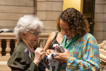 Vereadora Luciana Boiteux recebeu com encanto o broche da campanha Nem Presa Nem Morta (Foto oficial) Uma mulher branca de cabelos brancos curtos presenteia a outra com objetos pequenas e as duas olham atentas para os objetos em suas mãos.