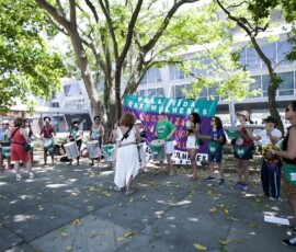 Mulheres dispostas em um semi-círculo tocam instrumentos de batucada na sombra de uma árvore e em frente a uma bandeira roxa e verde