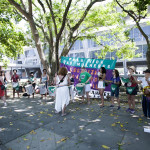 Plano geral da Batucada Unificada Foto: Kati Tortorelli. Mulheres dispostas em um semi-círculo tocam instrumentos de batucada na sombra de uma árvore e em frente a uma bandeira roxa e verde