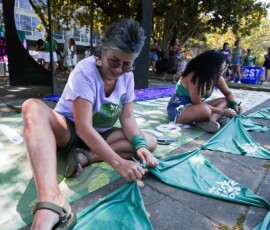 Duas mulheres sentadas no chão amarram lenços verdes uns nos outros.