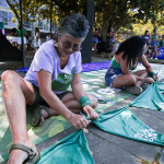 No Esquenta do dia 24, foi confeccionada a bandeira a ser pendurada nos Arcos da Lapa, na madrugada do Dia 28 de Setembro. Foto: Kati Tortorelli. Duas mulheres sentadas no chão amarram lenços verdes uns nos outros.