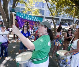 Diversas mulheres tocam instrumentos e batucada. Atrás há uma bandeira em roxo e verde.