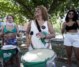 Três mulheres tocam instrumentos de batucada com lenços verdes.