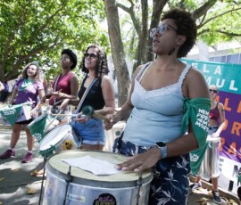 Mulher negra toca surdo em frente a uma bandeira com dizeres sobre aborto. Ela usa um lenço verde amarrado no braço.