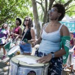 Batucada Unificada feminista no ensaio para a apresentação do Dia 28 de Setembro. Foto: Kati Tortorelli Mulher negra toca surdo em frente a uma bandeira com dizeres sobre aborto. Ela usa um lenço verde amarrado no braço.
