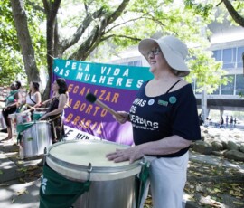 Mulher branca, com camiseta preta e chapéu branco, toca surdo em frete a uma bandeira verde e roxa onde se lê: Pela Vida das mulheres. Legalizar o aborto no Brasil.