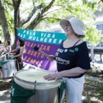 Batucada Unificada feminista no ensaio para a apresentação do Dia 28 de Setembro. Foto: Kati Tortorelli Mulher branca, com camiseta preta e chapéu branco, toca surdo em frete a uma bandeira verde e roxa onde se lê: Pela Vida das mulheres. Legalizar o aborto no Brasil.