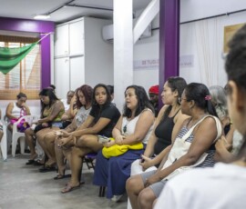 Diversas mulheres sentadas em cadeiras dialogam em uma sala com paredes brancas. Há um lenço verde grande ao fundo.