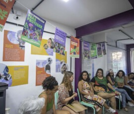 Algumas mulheres formam um semi-círculo sentadas em cadeiras. Há um varal com cartazes da Campanha Nem Presa Nem Morta.