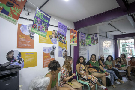 Bibiana Serpa fez a apresentação em nome da campanha Nem Presa Nem Morta. Foto: Gabi Lino. Algumas mulheres formam um semi-círculo sentadas em cadeiras. Há um varal com cartazes da Campanha Nem Presa Nem Morta.
