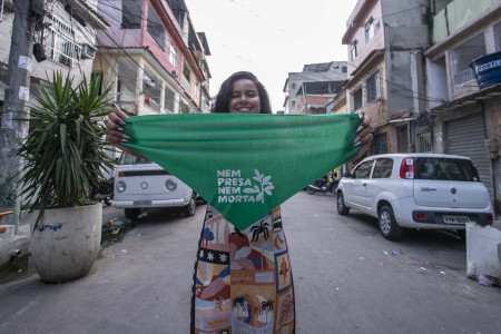 Cena na Maré, no dia da Roda de Conversa. Foto: Gabi Lino Mulher segura lenço verde da Nem Presa Nem Morta no meio da rua no Complexo da Maré.