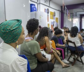 Visão de costas de mulheres sentadas em cadeiras, olhando para o centro e uma roda, onde falam as palestrantes.